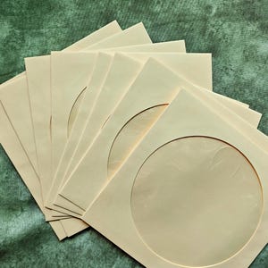 May include: A stack of cream-colored, square photo mats with circular openings. The mats are arranged in a fan shape, revealing a glimpse of the background. The background is a textured, dark green surface.