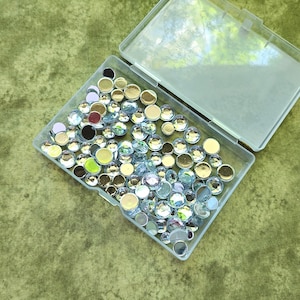 May include: A clear plastic container filled with assorted rhinestones. The rhinestones are various colors, including clear, gold, and iridescent. The container is open, revealing the contents. The background is a textured green fabric.