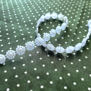 May include: A length of white floral lace trim with a pearl-like bead in the center of each flower. The lace is arranged in a curved shape against a green fabric background with white polka dots.