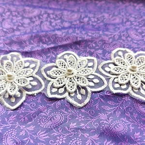 May include: Three white lace flower appliques with pearl centers on a purple background.