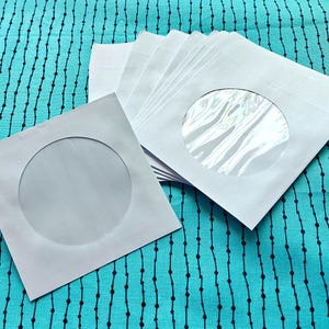 May include: A stack of white square envelopes with a circular window. The envelopes are on a teal fabric with a black dotted line pattern. The window reveals a clear, transparent material.
