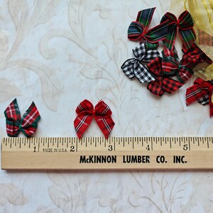 May include: Various miniature plaid bows in red, green, black, and white. A wooden ruler with inch markings and the text "MADE IN U.S.A." and "McKINNON LUMBER CO. INC." is in the foreground. The bows are scattered on a patterned surface.