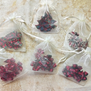 May include: Five sheer white organza drawstring bags, each containing a collection of small fabric bows. The bows feature various plaid patterns in red, green, and dark blue color schemes. The bags are arranged in a cluster on a textured surface.