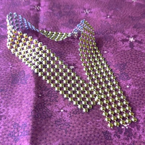 May include: A gold and silver chain necklace made of small, round beads. The chain is laid out in a curved shape on a purple fabric background.