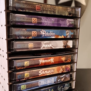 May include: A wooden organiser with eight slots for storing dice and game cards. Each slot features a different character class, including Treant, Shadow Thief, Paladin, Monk, Pyromancer, Samurai, Gunslinger, and Ninja. The organiser is made of wood and has a clear plastic front.