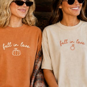 May include: Two short-sleeve crewneck t-shirts. The left shirt is burnt orange with white text reading fall in love above a pumpkin graphic. The right shirt is cream with rust-colored text reading fall in love above a ring graphic.