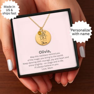 Op de afbeelding: Een goudkleurige ketting met een ronde hanger met balletschoenen en de naam "Olivia". De ketting heeft een kleine, gele edelsteen en wordt gepresenteerd in een zwarte geschenkdoos met een boodschap.
