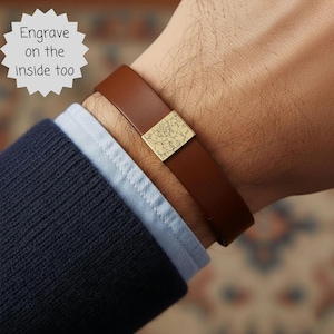 May include: A brown leather bracelet with a gold-colored square centerpiece featuring a map design. The bracelet is worn over a blue dress shirt and navy blue sweater. Text in the image reads, "Engrave on the inside too."