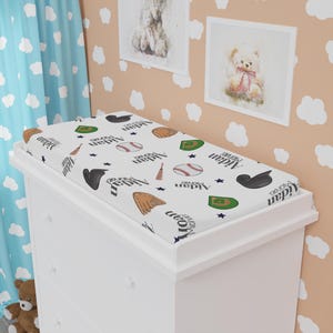 May include: A white changing pad cover with a baseball theme. The cover features baseballs, gloves, hats, and baseball field graphics. The name "Aidan Michael" is printed on the cover in a repeating pattern. The cover is on a white dresser.