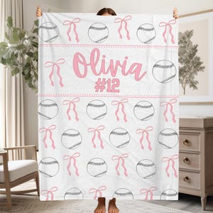 May include: White blanket with a baseball design. The pattern includes baseballs and pink bows. The name "Olivia" and the number "12" are displayed in pink. This blanket would be suitable for a child's bedroom or as a sports-themed present.