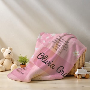 Op de afbeelding: Een roze babydeken met een gepersonaliseerde naam, Olivia Grace, en bijbelverzen. De deken heeft een patchwork-ontwerp met sterren, bloemen en een zon. Een teddybeer en een speelgoedauto staan op de achtergrond.