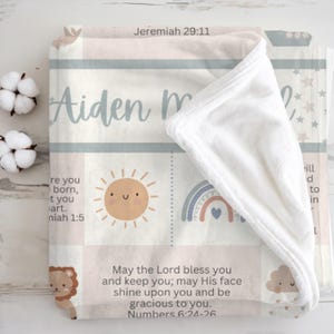May include: A baby blanket with a soft, plush texture. The blanket features a pastel color scheme with a sun, rainbow, and lion graphic. The name "Aiden m" is displayed, along with Bible verses. The blanket is folded over, revealing a white interior.