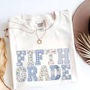 May include: A cream-coloured t-shirt featuring the text FIFTH GRADE in large block letters. Each letter has a unique blue and white pattern, including daisies, gingham checks, small floral prints, and bows. A gold-toned necklace rests on the shirt.