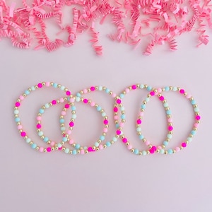 May include: Four beaded bracelets in pastel colors of pink, blue, and gold. The bracelets are arranged on a light pink surface, with pink paper shreds in the background. The bracelets are made of small beads.