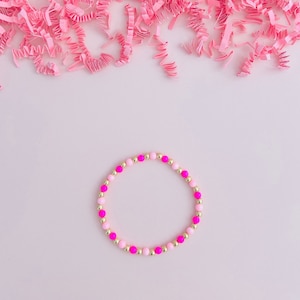 May include: A beaded bracelet featuring alternating pink and gold-colored beads. The bracelet is composed of small, round beads in shades of light pink, hot pink, and gold. The bracelet is displayed on a light pink surface with pink paper shreds.