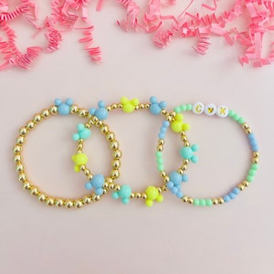 May include: Three beaded bracelets with gold-colored beads and colorful Mickey Mouse-shaped accents. One bracelet has the letters "CXK" on it. The bracelets are in shades of blue, green, and yellow.