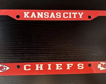 May include: A rectangular red license plate frame featuring the white text KANSAS CITY at the top and CHIEFS at the bottom. The frame includes two white arrowhead logos with red KC lettering on the bottom left and right corners of the frame.