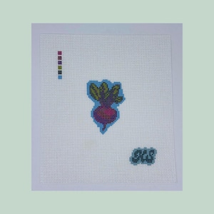 May include: A cross-stitch design featuring a purple beet with green leaves, outlined in blue, on a white background. The design includes a color palette and the letters "SCS" in blue.