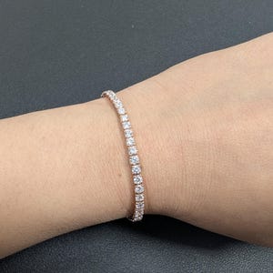 Puede incluir: Una pulsera de tenis de oro rosa con una fila de piedras preciosas claras y brillantes. La pulsera se lleva en la muñeca, mostrando el delicado diseño y el brillo de las piedras. Una pieza de joyería clásica.
