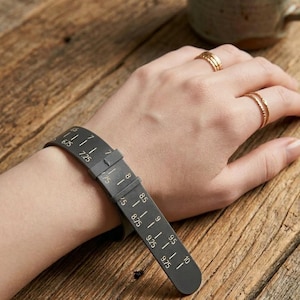 May include: A black wrist sizer tool with white markings, measuring from 6.75 to 10 inches. The tool is worn on a wrist, with a buckle closure. The background is a wooden surface.