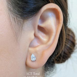 May include: A close-up of an ear with a pear-shaped diamond stud earring. The earring is set in a silver or white metal. The text below the earring reads "1CT Total (0.50CT Each)".