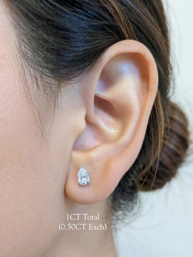 May include: A close-up of an ear with a pear-shaped diamond stud earring. The earring is set in a silver or white metal. The text below the earring reads "1CT Total (0.50CT Each)".