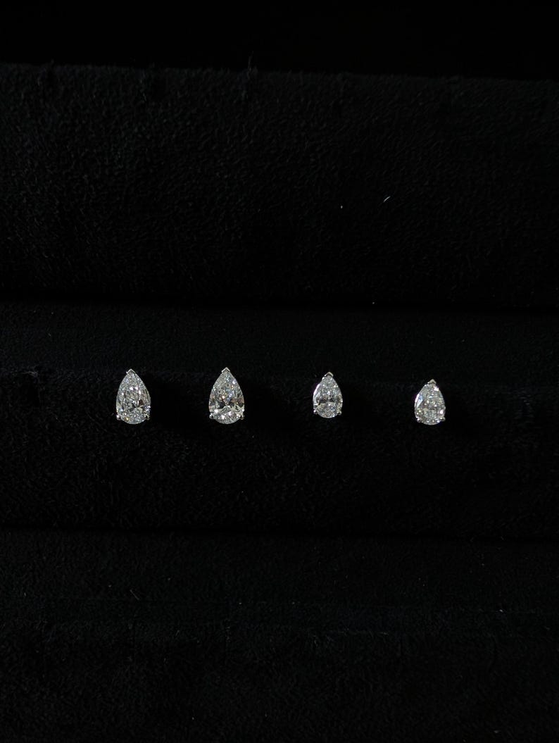 May include: Four pear-shaped diamond stud earrings of varying sizes are displayed against a black background. The diamonds sparkle, showcasing their facets. The earrings are arranged in a row, highlighting their graduated sizes.