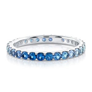May include: A silver ring with a band of blue gemstones. The gemstones graduate in color from light to dark blue. The ring is set against a white background.