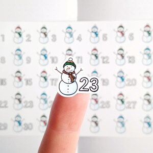 May include: A sheet of snowman stickers, each with a different number. One sticker, held by a finger, features a snowman wearing a green hat and brown scarf, with the number 23. The other stickers have snowmen with different colored hats and scarves.