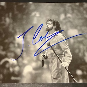 May include: A black and white photograph of a performer with dreadlocks and a beard, wearing a jumpsuit and holding a microphone on stage. The image features a prominent blue ink autograph across the centre and a small authentication sticker.