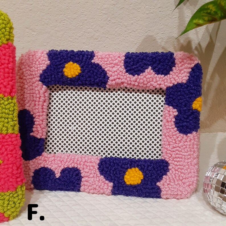 May include: A pink and purple floral picture frame with a white mesh insert. The frame is made of yarn and has a textured, fuzzy appearance.