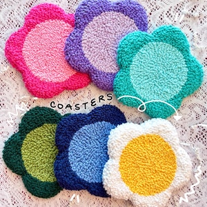 May include: A collection of handmade, flower-shaped coasters in various colors. The coasters are made of textured yarn and include shades of pink, purple, teal, green, blue, white, and yellow. The word "COASTERS" is printed on the image.