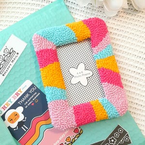 May include: A colorful 4x6 inch photo frame made with yarn and a rainbow color scheme. The frame is pink, yellow, blue, and turquoise.