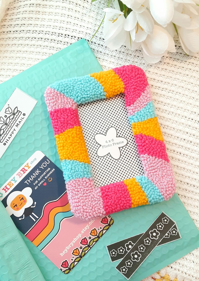May include: A colorful 4x6 inch photo frame made with yarn and a rainbow color scheme. The frame is pink, yellow, blue, and turquoise.