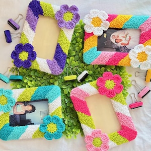 May include: Four rectangular picture frames featuring textured, diagonal stripes in vibrant colors like purple, lime green, white, pink, yellow, and turquoise. Each frame is adorned with two large, crocheted flower accents in contrasting colors.