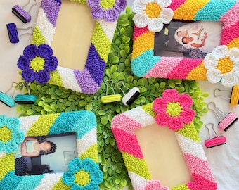 May include: Four rectangular picture frames featuring textured, diagonal stripes in vibrant colors like purple, lime green, white, pink, yellow, and turquoise. Each frame is adorned with two large, crocheted flower accents in contrasting colors.