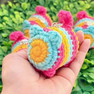 May include: A small, cylindrical crochet plush toy featuring horizontal stripes in vibrant shades of hot pink, bright yellow, and light blue. The ends of the cylinder are finished with a spiral pattern in yellow and blue, and a small metal ring is attached.