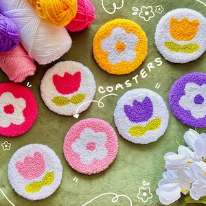 May include: A collection of handmade, round coasters with floral designs. Each coaster features a white background with a colorful flower or tulip design in shades of pink, yellow, purple, and orange. The word "COASTERS" is written in white script.