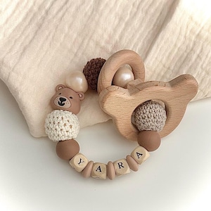 Könnte beinhalten: Ein Baby-Beißring mit einem hölzernen bärenförmigen Ring, einer braunen gehäkelten Perle, einer cremefarbenen gehäkelten Perle, glatten braunen und cremefarbenen Perlen sowie Holzbuchstabenperlen, die YARA bilden, ergänzt durch kleine Silikonperlen.
