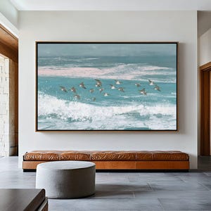 Könnte beinhalten: Große gerahmte Kunstwerk, das eine Küstenlandschaft mit Vögeln zeigt, die über den Meereswellen fliegen. Das Kunstwerk verwendet Blau-, Weiß- und Rosatöne mit einem schwarzen Rahmen. Das Kunstwerk wird über einer braunen Lederbank ausgestellt.