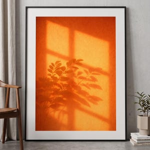 Op de afbeelding: Een ingelijste kunstdruk met een oranje muur met de schaduw van een raam en een bladplant. De schaduwen creëren een opvallend contrast met de textuur achtergrond. De print zit in een zwart frame en wordt getoond in een kamer met een stoel en een potplant.