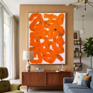 May include: Large abstract painting with bold orange brushstrokes on a white background, framed in black. The artwork hangs above a mid-century modern wooden cabinet. A lamp with a patterned base and a white shade sits on the cabinet.