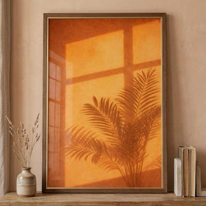 Op de afbeelding: Ingelijste kunstdruk met een warme oranje achtergrond, met de schaduw van een palmboom en een raam. De lijst is donkerbruin met een gouden binnenrand. De print wordt gepresenteerd op een houten plank, wat een warme sfeer creëert.