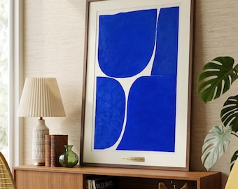 May include: Framed abstract art print featuring bold blue shapes against a white background. The artwork is displayed in a dark wood frame and is mounted on a neutral-toned wall. A lamp, books, and a green vase are on a wooden cabinet.
