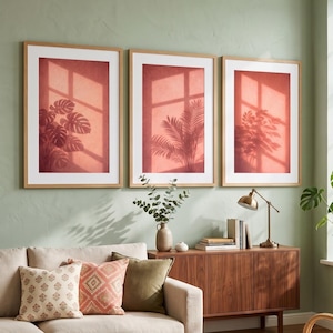 May include: A set of three vertical wall art prints in light wood frames, each featuring a terracotta-colored background with a white window pane shadow and the dark silhouette of a different tropical plant, including monstera and palm leaves.