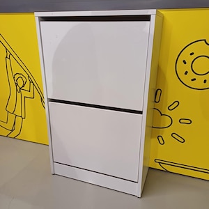 May include: White two-tiered shoe cabinet with two doors. The cabinet has a simple design and is made of wood.