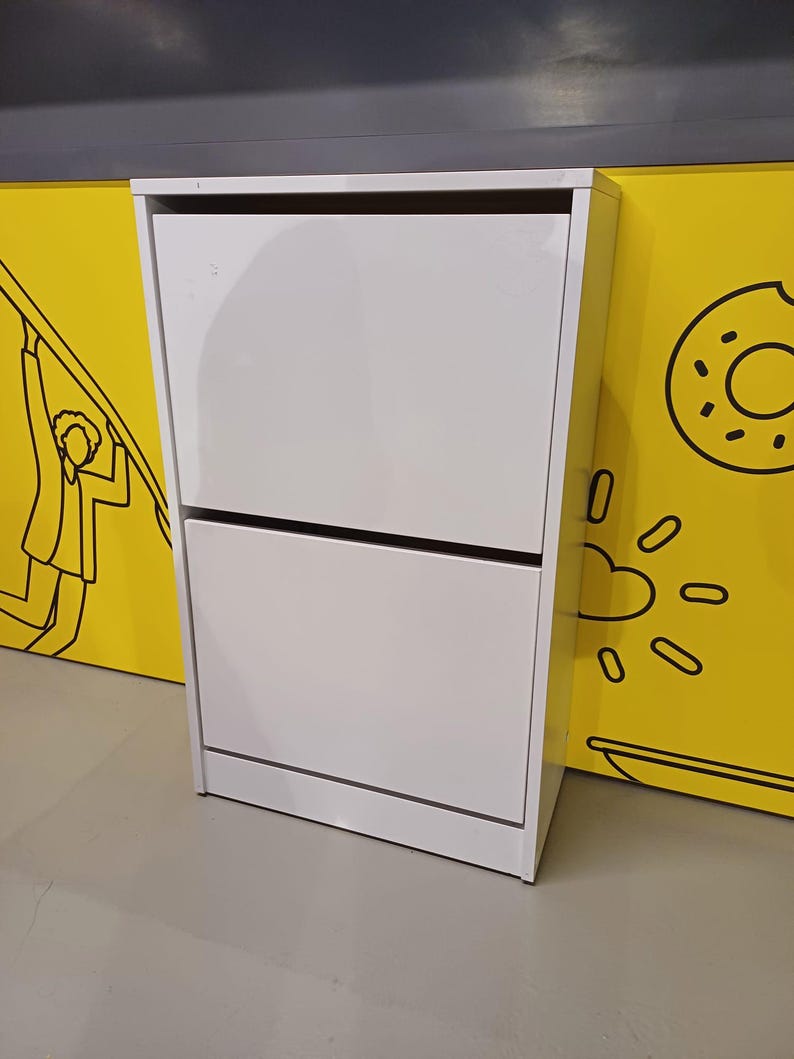 May include: White two-tiered shoe cabinet with two doors. The cabinet has a simple design and is made of wood.