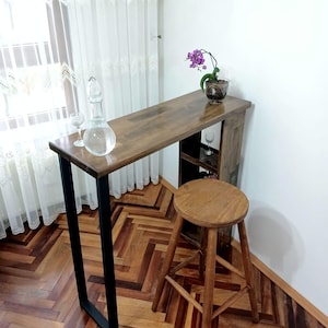 May include: A wooden bar table with a black metal frame and a wooden stool. The table has a shelf and a small plant in a pot on top. The table and stool are in a room with a wooden floor.