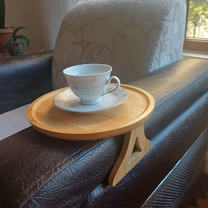 Könnte beinhalten: Ein helles Sofa-Armtablett aus Holz mit einer Tasse und Untertasse. Das Tablett hat eine runde Oberseite und eine dreieckige Stütze. Die Tasse und Untertasse sind weiß mit einem zarten Blumenmuster. Der Sofararm ist dunkelbraun.