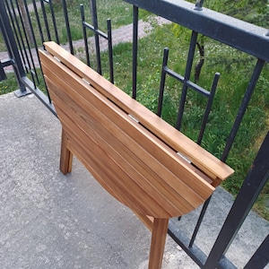 May include: A wooden folding table with a rounded top and a natural wood finish. The table is folded and standing on a concrete surface.